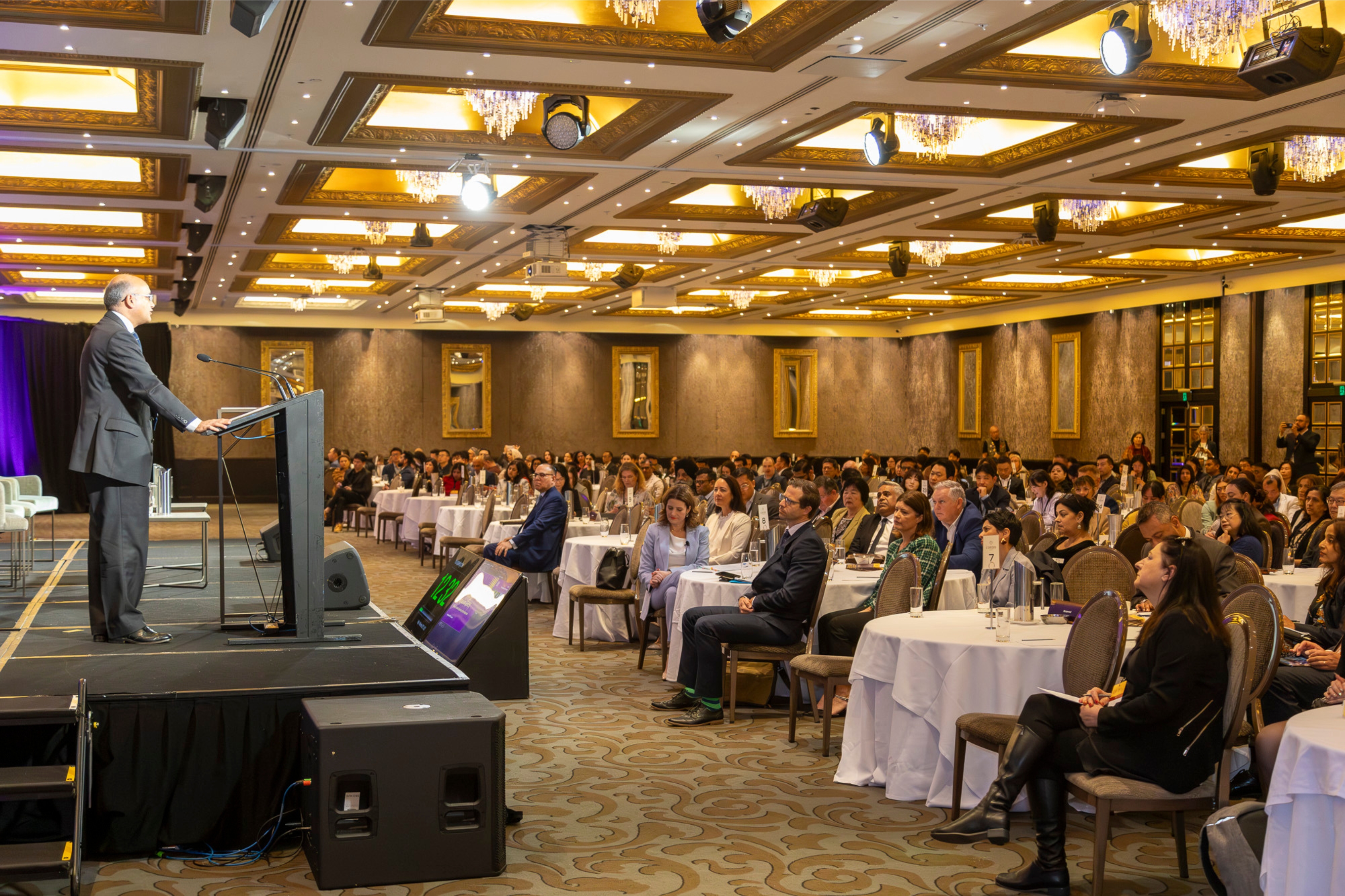 A person stands on stage, speaking into a microphone. A large seated audience is seen in the background.