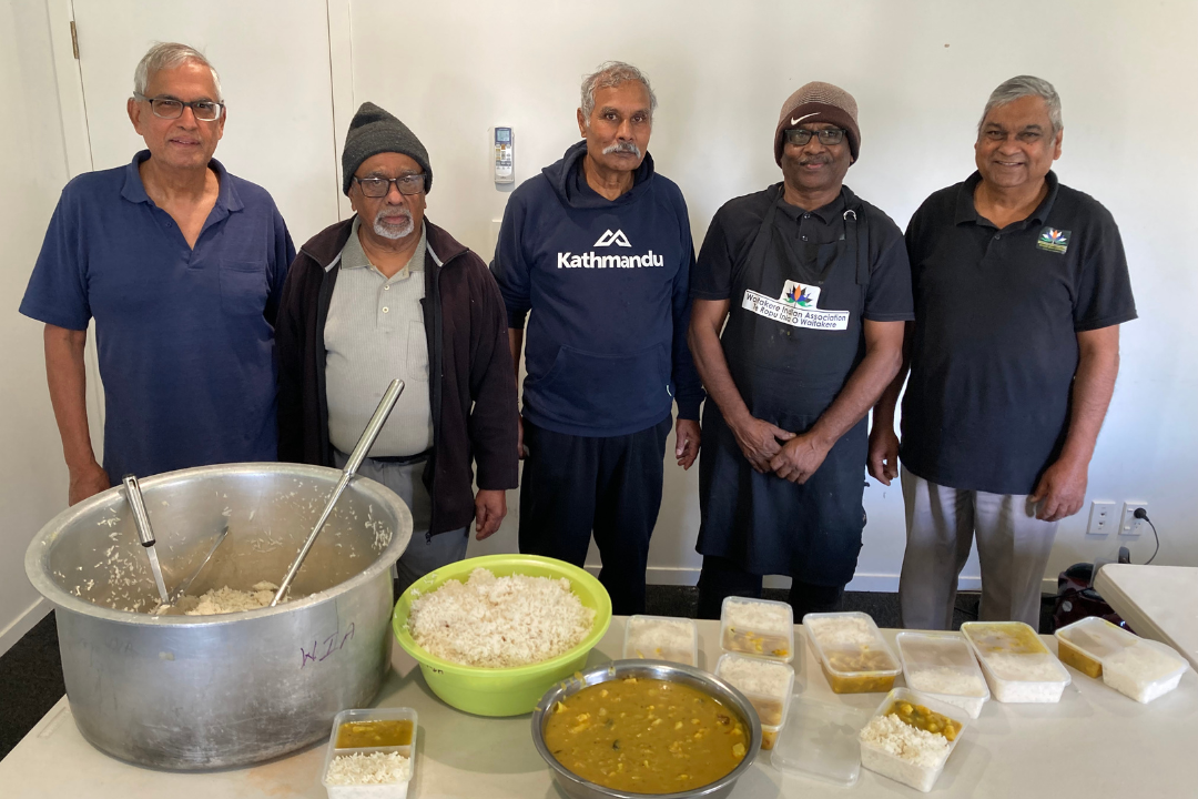 Five people stand, posing for a photo in front of a table of food, including large posts of rice and dahl and packaged meals