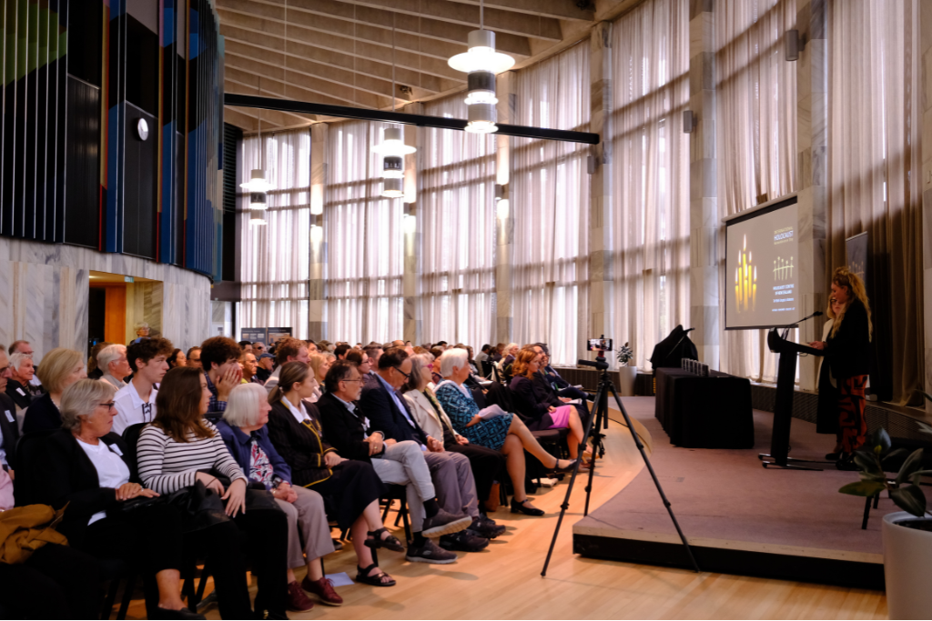 A speaker addresses a large seated audience in a modern hall with tall sheer curtains and overhead lighting. A presentation slide is projected on a screen beside the speaker.