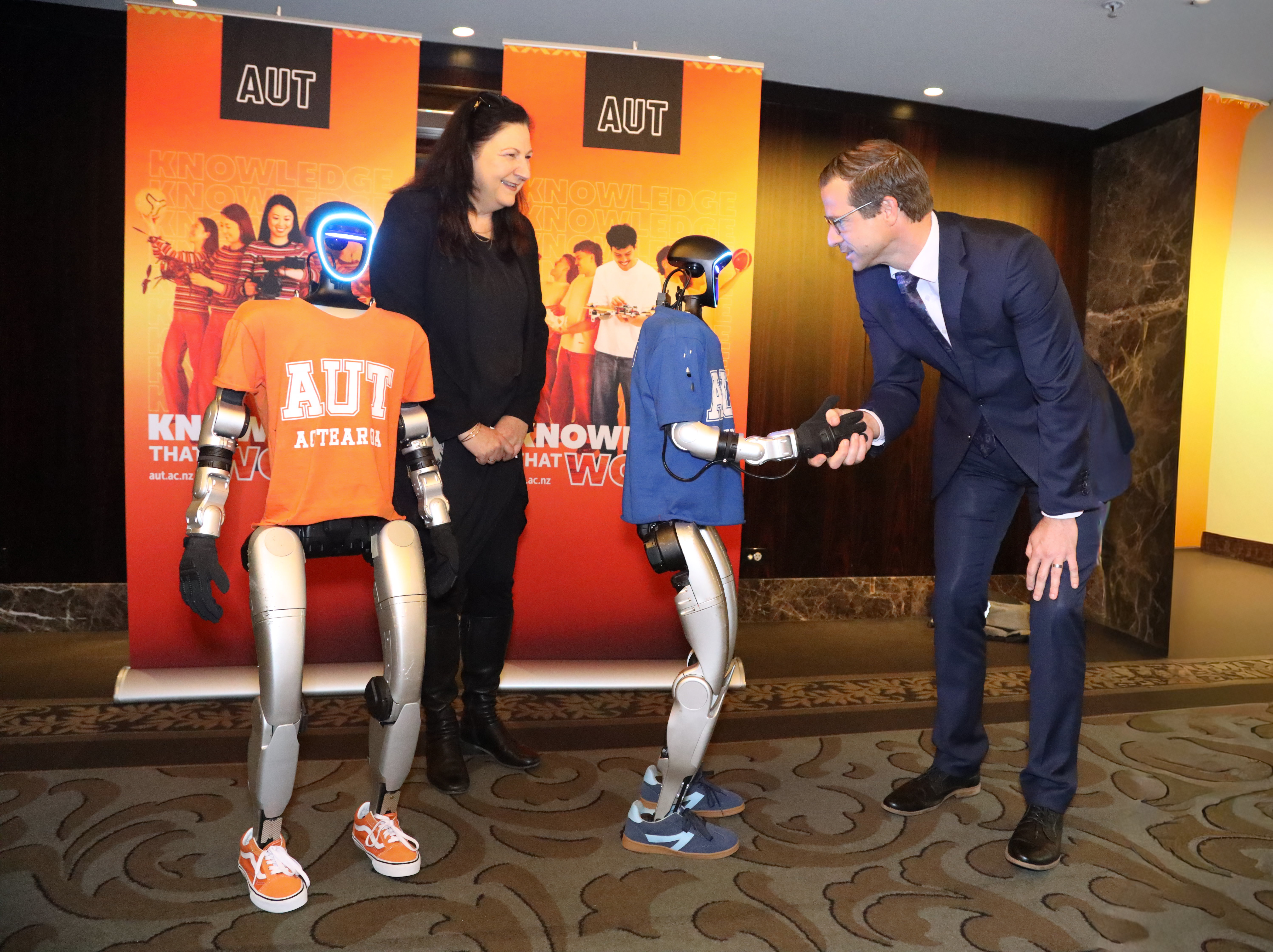 Two people stand with two humanoid robots, one of them shaking hands with one of the humans.