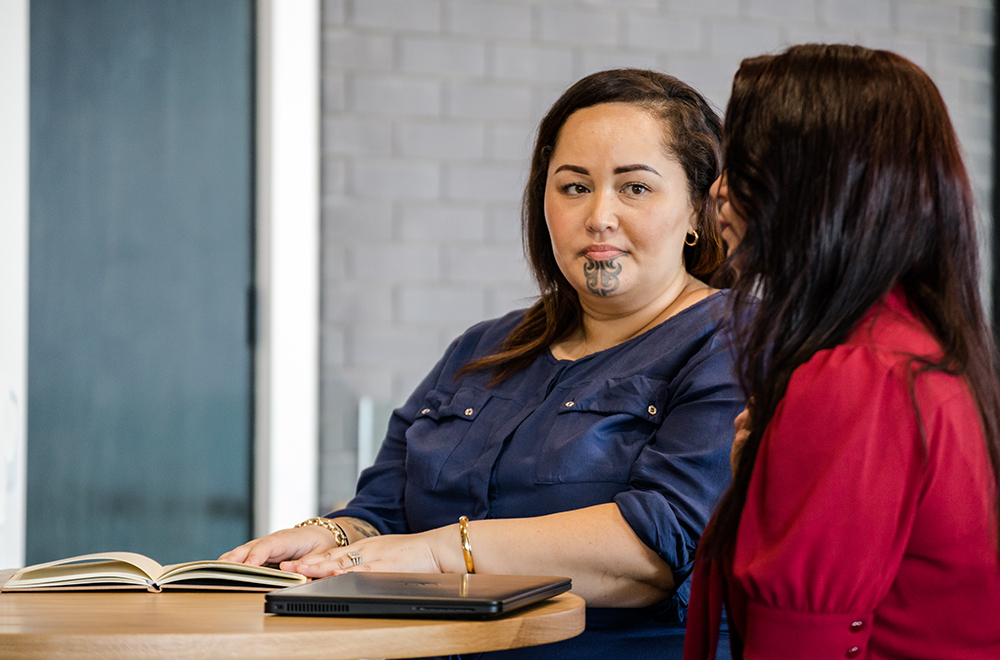 Image of 2 women sitting at a table the person in the foreground is facing away. The other is facing forward towards the camera and she has a tā moko (traditional tattooing)