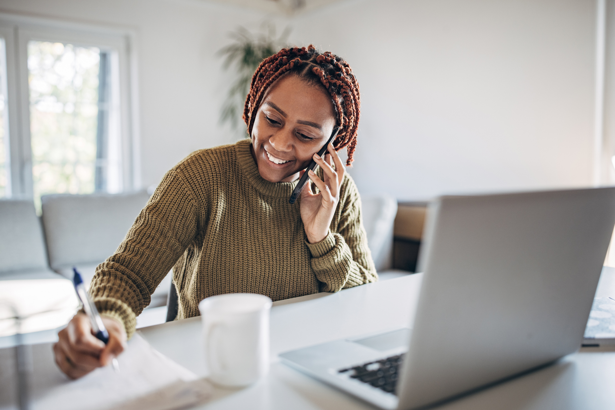 Image - woman sitting at a desk with pen and paper and a laptop open in front and a phone on her ear