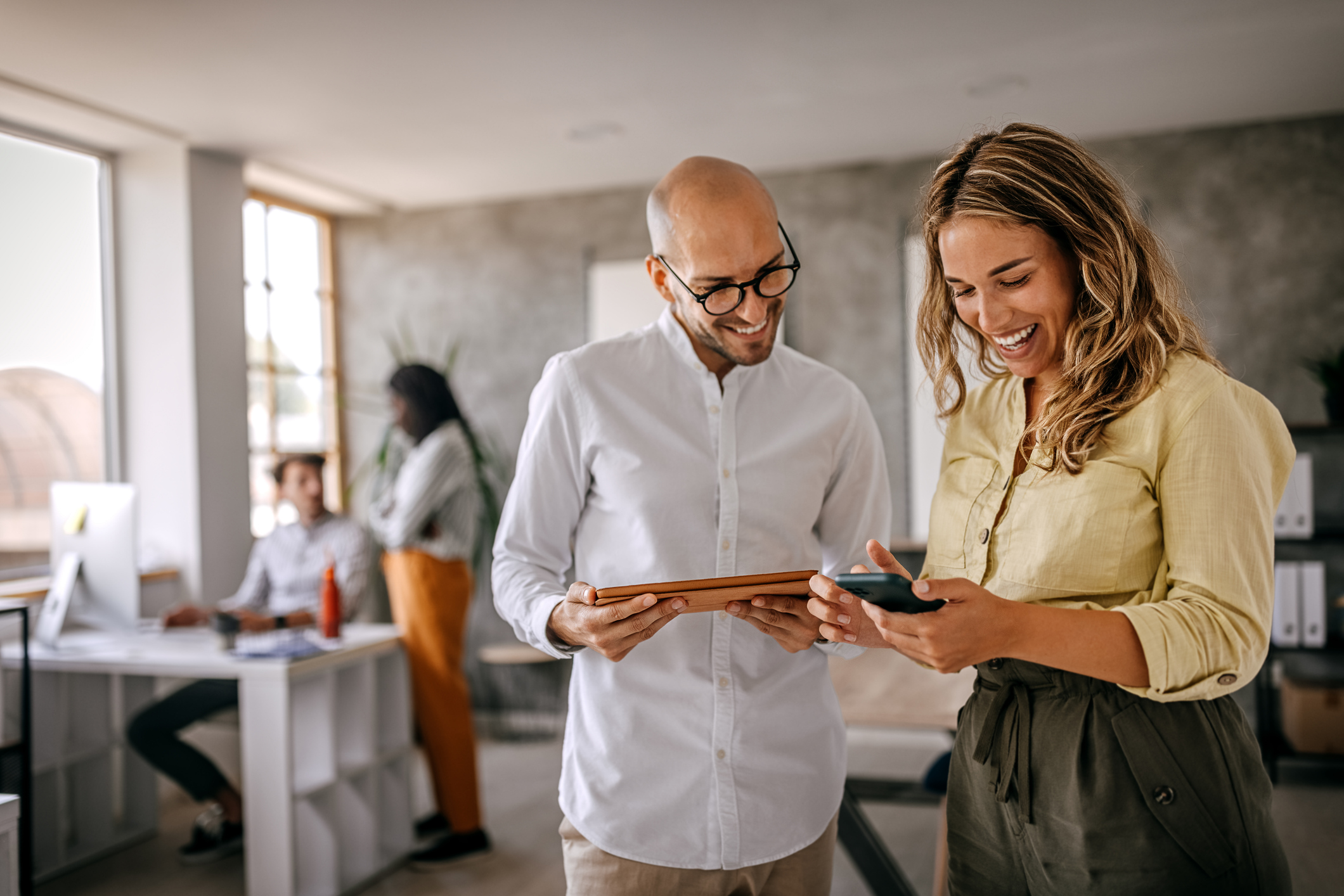 Image of an office with people looking over a phone in the foreground with a person sitting at a desk and somebody standing next to it in the background