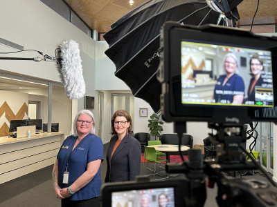 Two women stand in front of a microphone and camera. The camera screen features in the foreground.